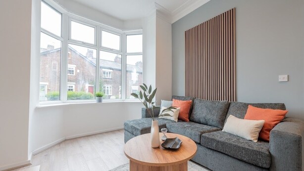 A modern living room with a grey sofa, a wooden coffee table, and a large window offering a view of buildings outside. - Comfort & Style in Salford - Sleeps 8 (Salford)