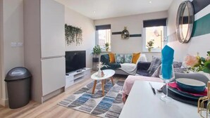 Een moderne woonkamer met een bank, salontafel en televisie. Op de vloer staat een afvalbak en een plant.