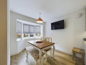 Een eetgedeelte met een houten tafel, witte stoelen, een bank en een aan de muur bevestigde televisie.