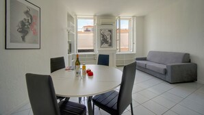 A dining area with a round table, chairs, a sofa, and two windows with a view of buildings. - Charmant 2 Pieces Proche Forville A1b269 (Cannes)