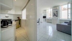 A modern kitchen with white cabinetry, a coffee machine, and a compact dishwasher. The living area features a grey sofa, a flat-screen TV, and a large window offering a view of the cityscape. - Charmant 2 Pieces Proche Forville A1b269 (Cannes)