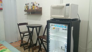 A small kitchenette with a microwave and a mini-fridge, a wooden chair, a small table, and a shelf with various bottles. - Cozy private suite with AC and Wi-Fi, just 3 minutes from a beautiful waterfall (Caraguatatuba)