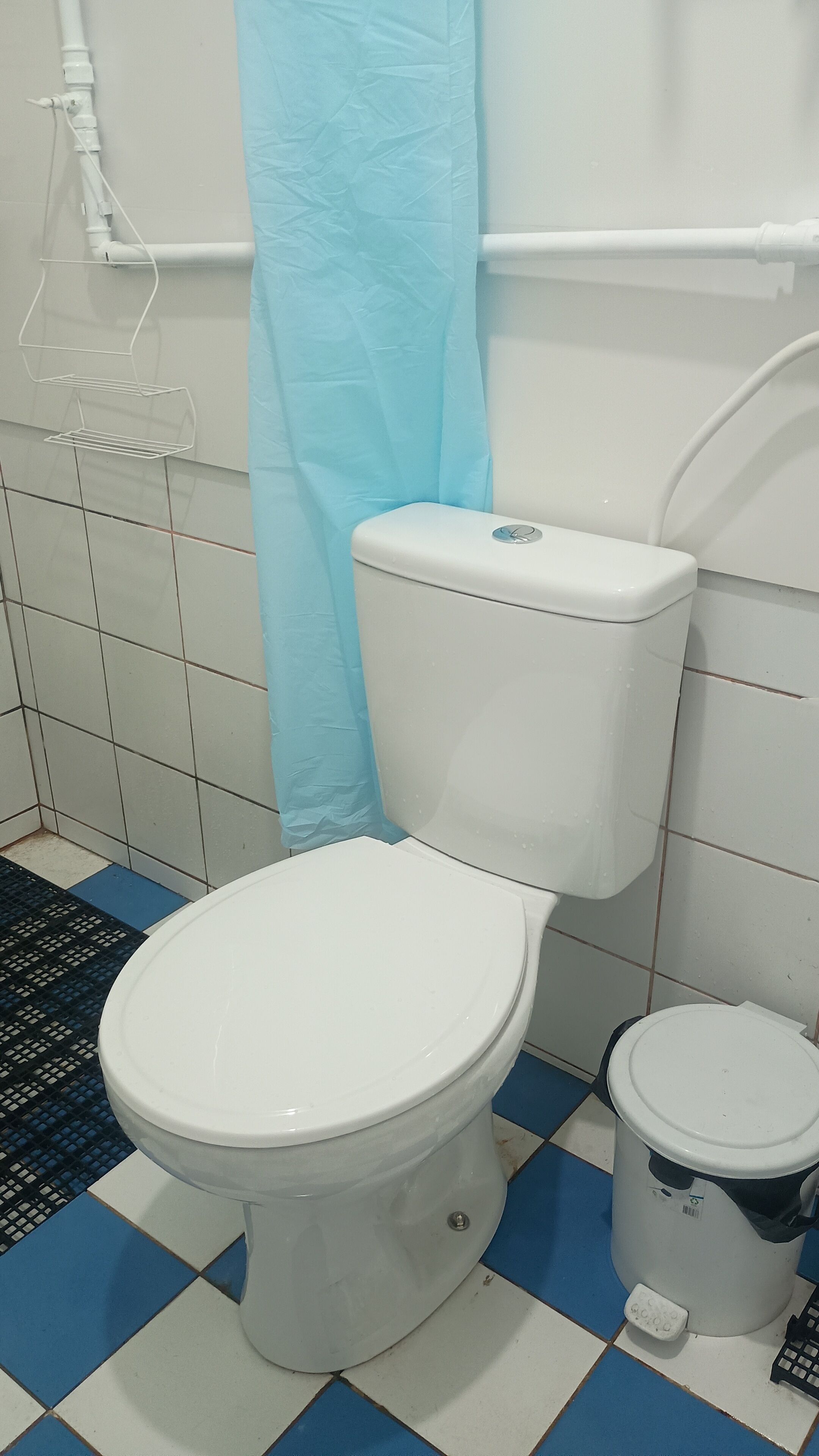 Une salle de bain avec des toilettes blanches, un bidet et une poubelle. Le sol est recouvert d’un motif à carreaux bleus et blancs.
