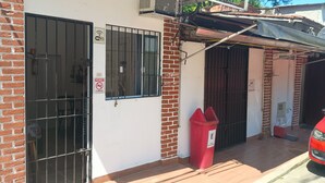 A building with a white exterior, a barred garage door, and a red trash bin. - Cozy private suite with AC and Wi-Fi, just 3 minutes from a beautiful waterfall (Caraguatatuba)