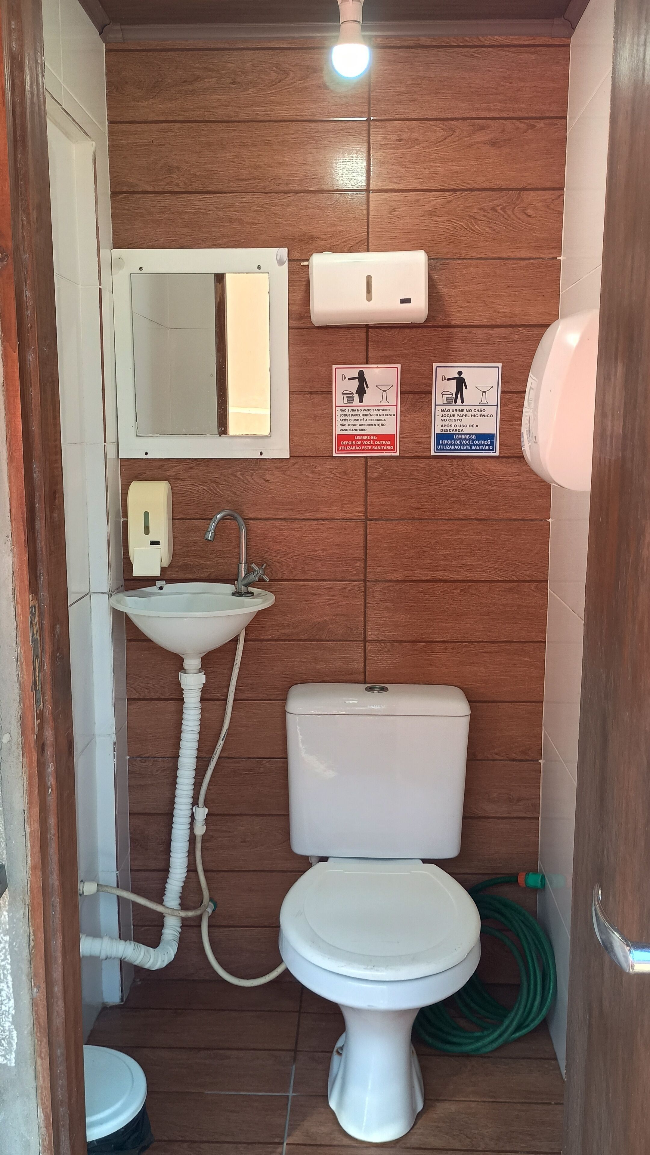 Casa de banho compacta com sanita, lavatório e espelho. Existem dois sinais na parede, um a indicar lavar as mãos e outro a indicar proibição de urinar.
