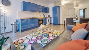 Una cocina moderna con armarios azules, un microondas y una lavadora. Hay una alfombra colorida en el suelo y un sofá con cojines naranjas en la sala.