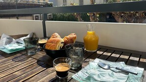 Een houten tafel met croissants, een glas sinaasappelsap en een kom koffie op een balkon met uitzicht op gebouwen.