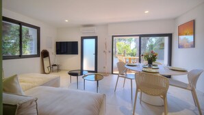 A modern living room with a sofa, a coffee table, and a dining area with chairs and a table set for two. There is a TV mounted on the wall, a door leading to an outdoor area, and a window with a view of greenery. - 2 Chambres Terrasse Jardin Acces Plages A2b241 (Cannes)