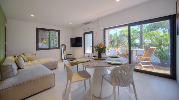 A modern living room with a sofa, dining table, and chairs, a flat-screen TV, and a large sliding glass door leading to an outdoor area with chairs and a table. - 2 Chambres Terrasse Jardin Acces Plages A2b241 (Cannes)