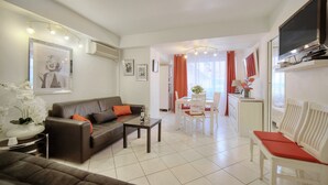 Un salon moderne avec un canapé, une table à manger et des chaises, un téléviseur fixé au mur, et un coin cuisine à l’arrière-plan.
