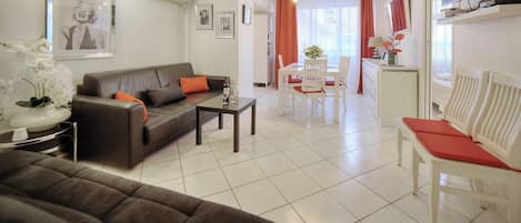 Una sala de estar moderna con sofá, mesa de comedor y sillas. Hay una zona de cocina con refrigerador y televisor.