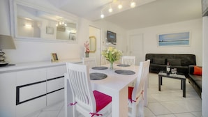 Une cuisine moderne avec des meubles blancs, une table à manger accompagnée de chaises, un téléviseur fixé au-dessus du comptoir et un canapé dans l’espace salon.