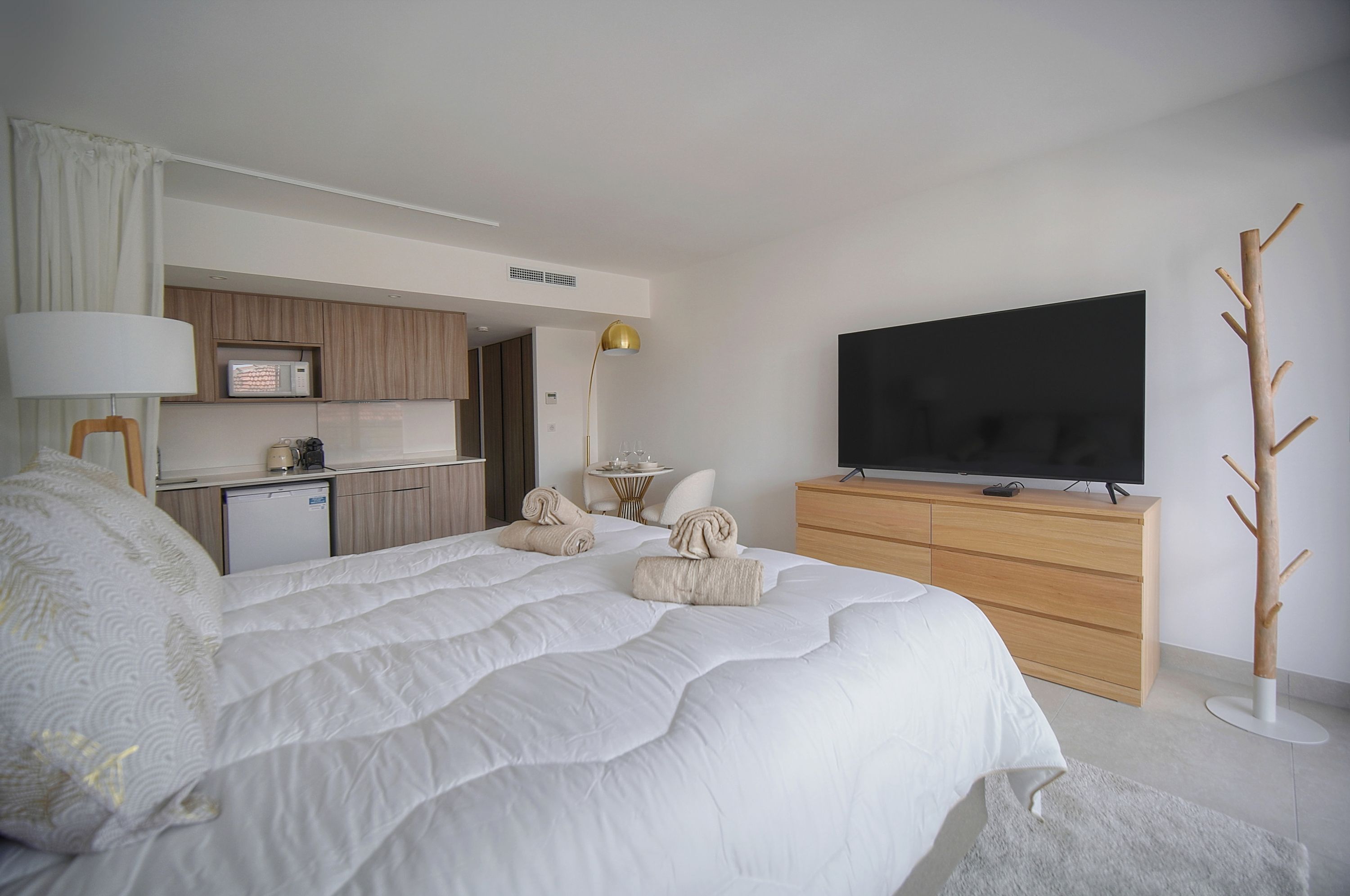 A modern hotel room with a large bed, a flat-screen TV, a wooden dresser, and a kitchenette in the background.