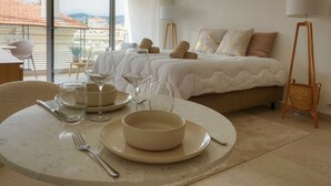 Une chambre d’hôtel avec un lit, des lampes de chevet, une table à manger avec des assiettes et des verres, et une vue sur la ville à travers de grandes fenêtres.
