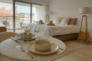 A hotel room with a bed, bedside lamps, a dining table with plates and glasses, and a view of the city through large windows. - Superbe Studio Neuf ET Moderne A0b196 (Cannes)
