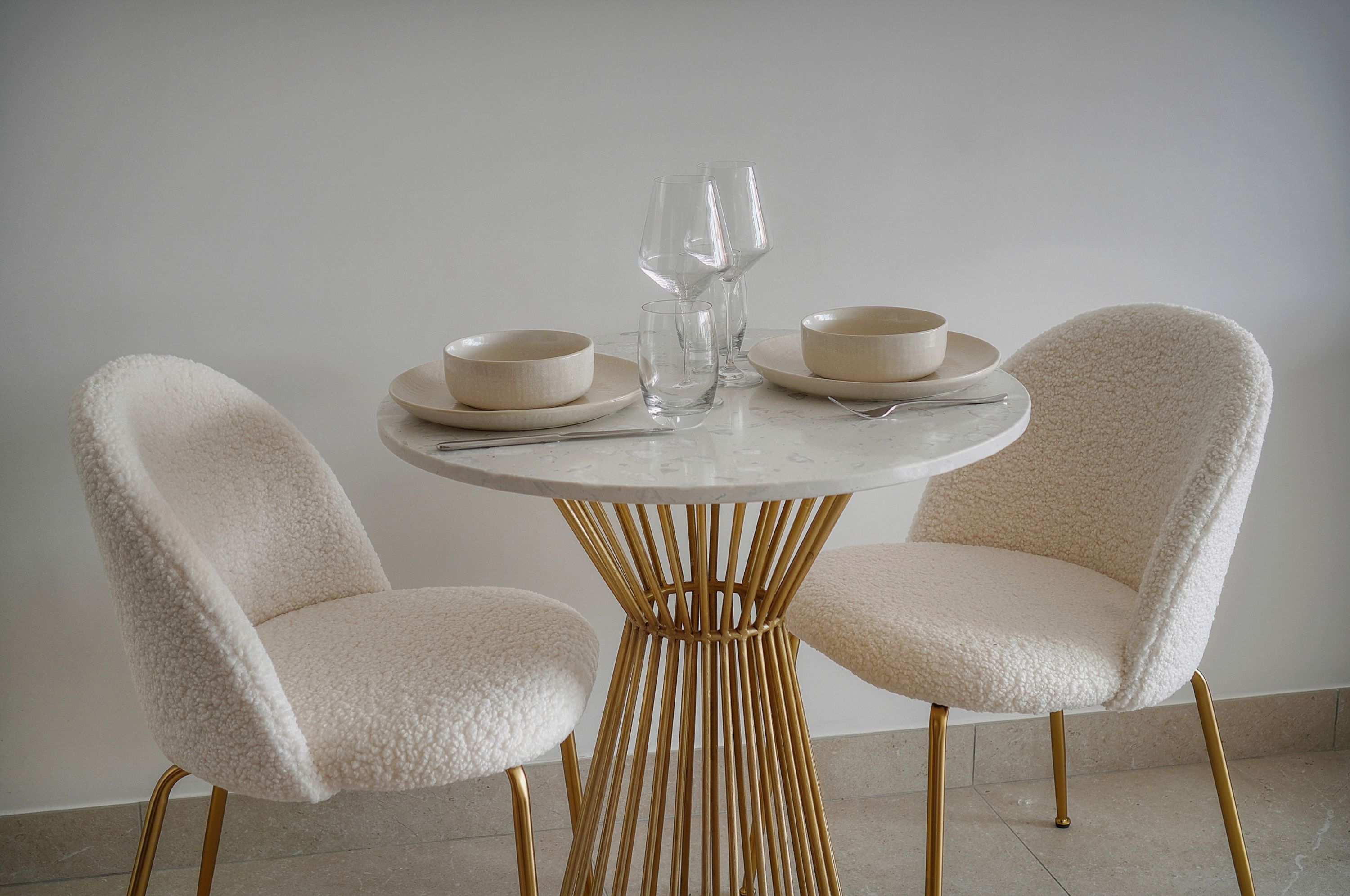 Une table ronde, accompagnée de deux chaises, chacune dotée d’un coussin à texture particulière, ainsi qu’une paire de verres à vin et de coupes placés sur la table.