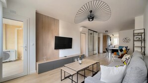 A modern living room with a flat-screen TV, a wooden slatted wall, a dining area with a table and chairs, and a gray sofa with a colorful pillow. - 3 Pieces Moderne Proche Martinez ET Plages A2b249 (Cannes)