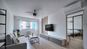 A modern living room with a grey sofa, a dining table, a TV mounted on the wall, and a large window with a view of buildings. - 3 Pieces Moderne Proche Martinez ET Plages A2b249 (Cannes)