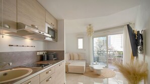 A modern kitchen with a built-in oven, microwave, and a large sink. There is a dining area with a white table and chairs, and a sofa. The room has a large sliding glass door leading to a balcony. - Superbe Studio Terrasse Parking A0b200 (Cannes)
