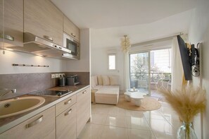 Eine moderne Küche mit eingebautem Backofen, Mikrowelle und einer großen Spüle. Es gibt eine Essecke mit einem weißen Tisch und Stühlen, sowie ein Sofa. Der Raum verfügt über eine große Schiebeglasdoor zur Balkon.