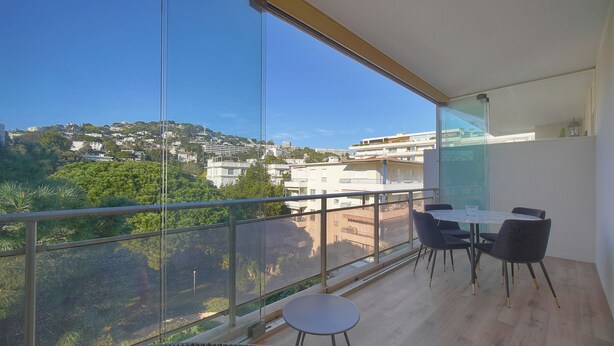 Een balkon met glazen hekwerk, een ronde tafel met stoelen, en een rieten stoel.