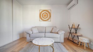 Een woonkamer met een grijze bank, een ronde salontafel met een marmeren blad, een kamerkleed met patroon en een kunstwerk aan de muur.