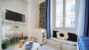 A modern living room with a flat-screen TV, a dining table with a bowl and a bottle, a sofa with a decorative pillow, and a window with a view of buildings. - Charmant Studio Hypercentre A0b211 (Cannes)