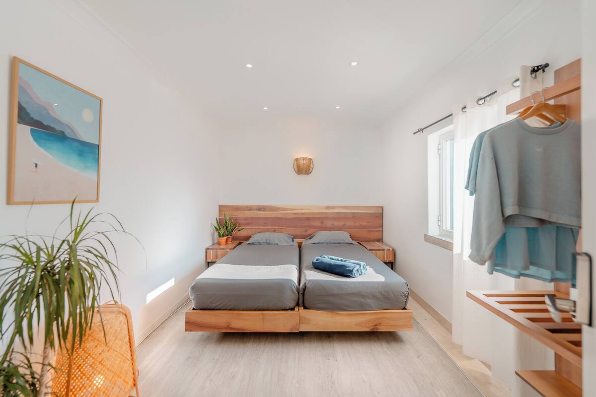 A bedroom with a wooden bed frame, two single beds, a bedside table, a hanging light fixture, a wall-mounted shelf, and a framed picture.