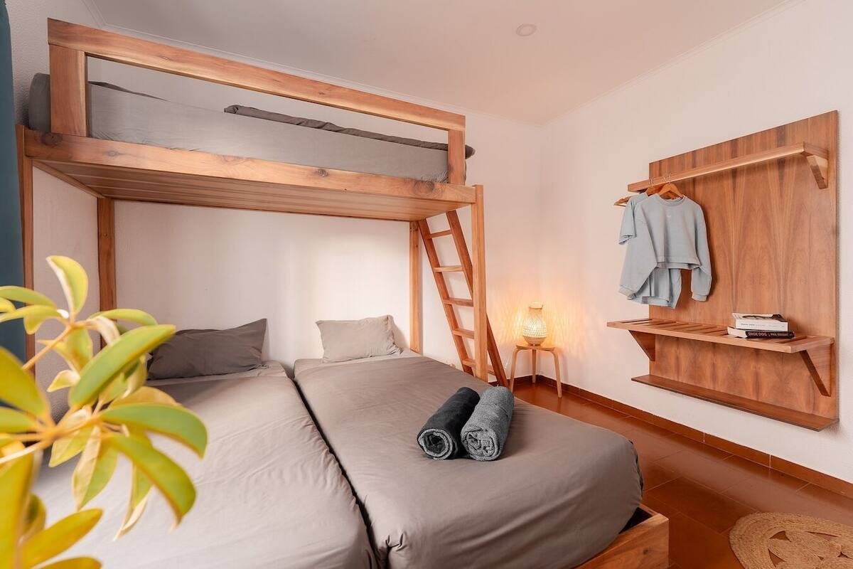 A bunk bed room with a ladder, a bedside table with a lamp, a shelf with books, and a hanging clothes rack.