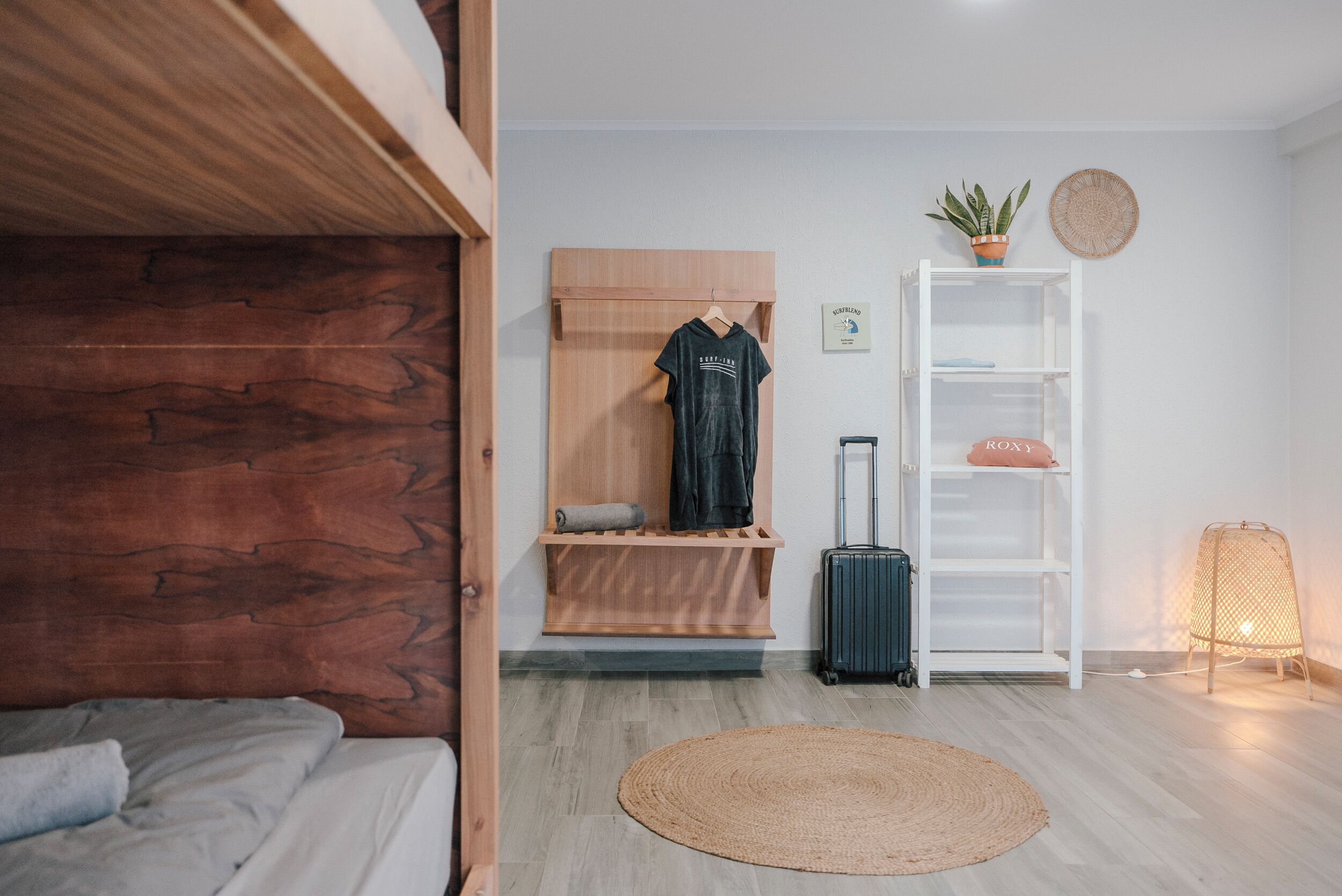 A room with a bunk bed, a wooden bench with hanging clothes, a suitcase, a white shelving unit, a woven rug, and a small potted plant.