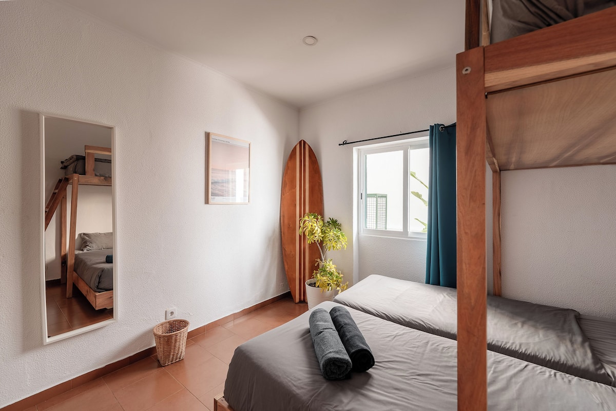A bedroom with a bunk bed, a single bed, a surfboard leaning against the wall, a framed picture, and a potted plant.