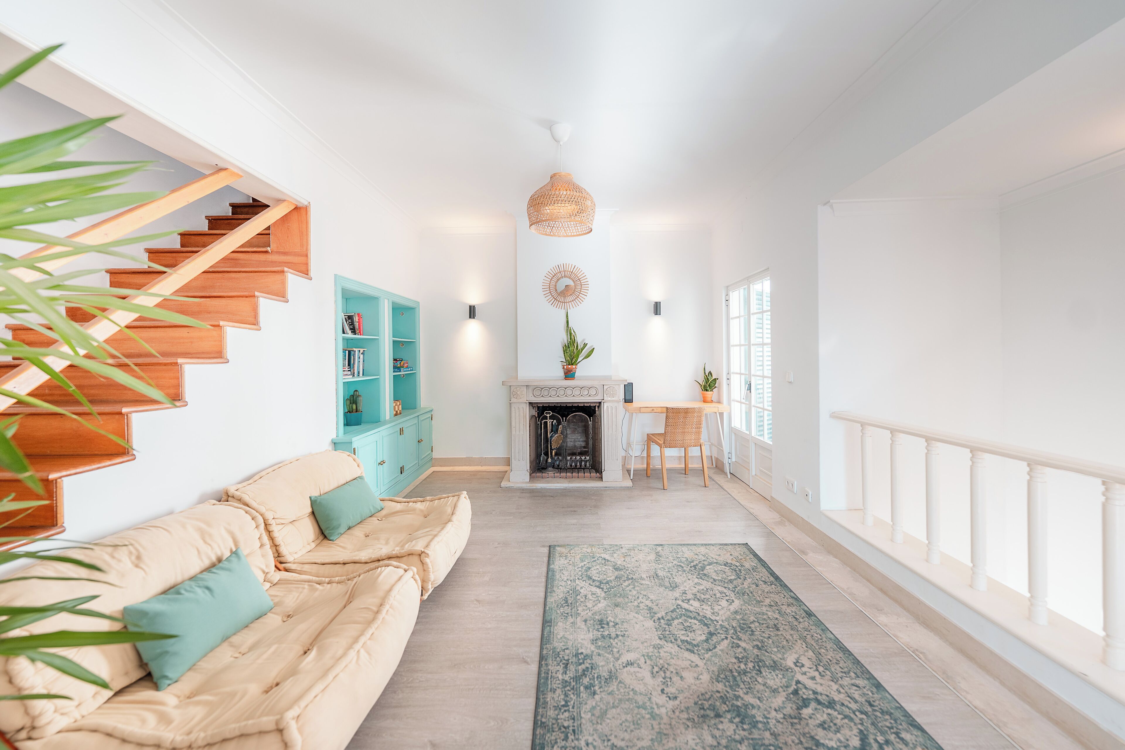Un salón espacioso con una escalera de madera, una chimenea, una estantería color turquesa y un amplio ventanal.