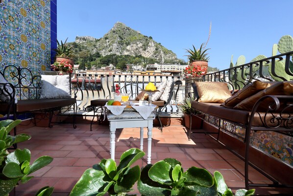 Una terraza con mesa y sillas blancas, además de un sofá. Hay plantas en macetas y se ve una montaña.