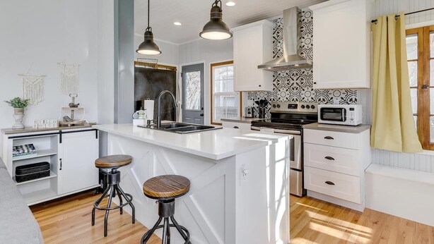 A modern kitchen with white cabinetry, a central island with stools, pendant lighting, and a large window with curtains. - Cozy North End Farmhouse  Easily Walk Downtown (Boise)