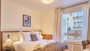 A hotel room with a large bed, a nightstand with a lamp, a framed picture on the wall, and a window with a view of buildings. - Sublim AC Apartment 3 Bdr Saint Germain des Prés (Paris)
