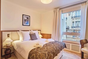 Ein Hotelzimmer mit einem großen Bett, einem Nachttisch mit Lampe, einem gerahmten Bild an der Wand und einem Fenster mit Blick auf Gebäude.