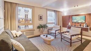 A modern living room with a sofa, armchairs, a coffee table, and a TV mounted on a wooden wall. Large windows offer views of a building with balconies. - Sublim AC Apartment 3 Bdr Saint Germain des Prés (Paris)