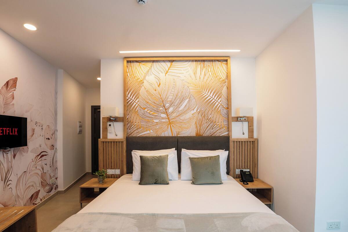 Une chambre d’hôtel moderne dotée d’un grand lit, de deux tables de chevet, d’un téléviseur fixé au mur et d’un mur décoratif orné d’un motif de feuilles.