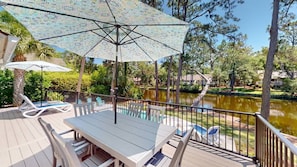 A deck with a table and chairs, a patterned umbrella, and a view of a lake. - Coastal Luxe Lagoon with Pool Near Beach (Hilton Head Island)