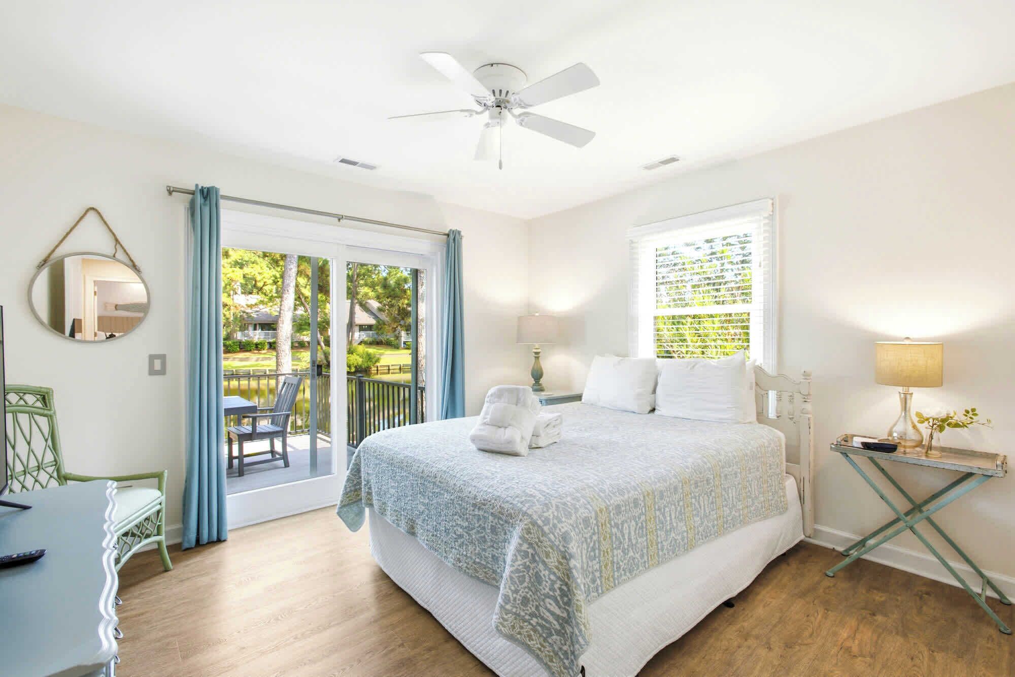 A bedroom with a bed, a ceiling fan, a window with curtains, a bedside table with a lamp, and a view of the outdoors.