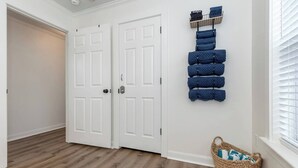 Un couloir avec deux portes blanches, un plancher en bois, une étagère murale où sont rangés des vêtements pliés, et un panier en osier près de la fenêtre.