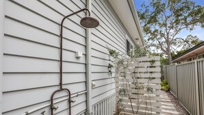 Une douche extérieure résidentielle, équipée d’un tuyau d’arrosage et d’un pommeau de douche à main, d’une clôture en bois et d’un bac à fleurs avec des plantes.