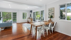 Un vaste espace de salle à manger avec une table en bois et des chaises blanches, de grandes fenêtres donnant sur un paysage verdoyant, et un canapé bleu en arrière-plan.