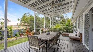 Une terrasse couverte avec une table et des chaises en bois, un banc rembourré et une vue sur un jardin et les maisons voisines.