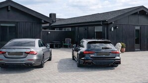 Two luxury cars parked in front of a modern building with large windows and a paved driveway. - 4 Star Holiday Home in Blokhus (Blokhus)
