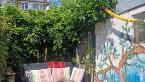 Ein Korbosofa mit bunten Kissen auf einer Holzterrasse, umgeben von üppiger Vegetation und einer Zierwand.