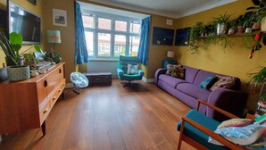 Ein Wohnzimmer mit Holzboden, einem violetten Sofa, einem blauen Sessel, einem Fernseher und einem Fenster mit Vorhängen.