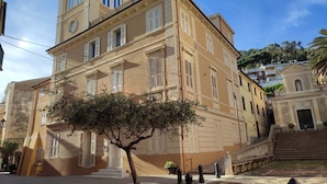 Um edifício histórico com uma torre central, várias janelas com persianas e uma escadaria que leva à entrada.
