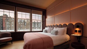 Habitación de hotel con una cama grande, una mesita de noche, una silla y vistas a edificios a través de amplias ventanas.