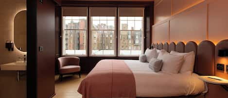 Un dormitorio con una cama grande, una silla, una mesita de noche, un ventanal con vistas a la ciudad y un baño con lavamanos.